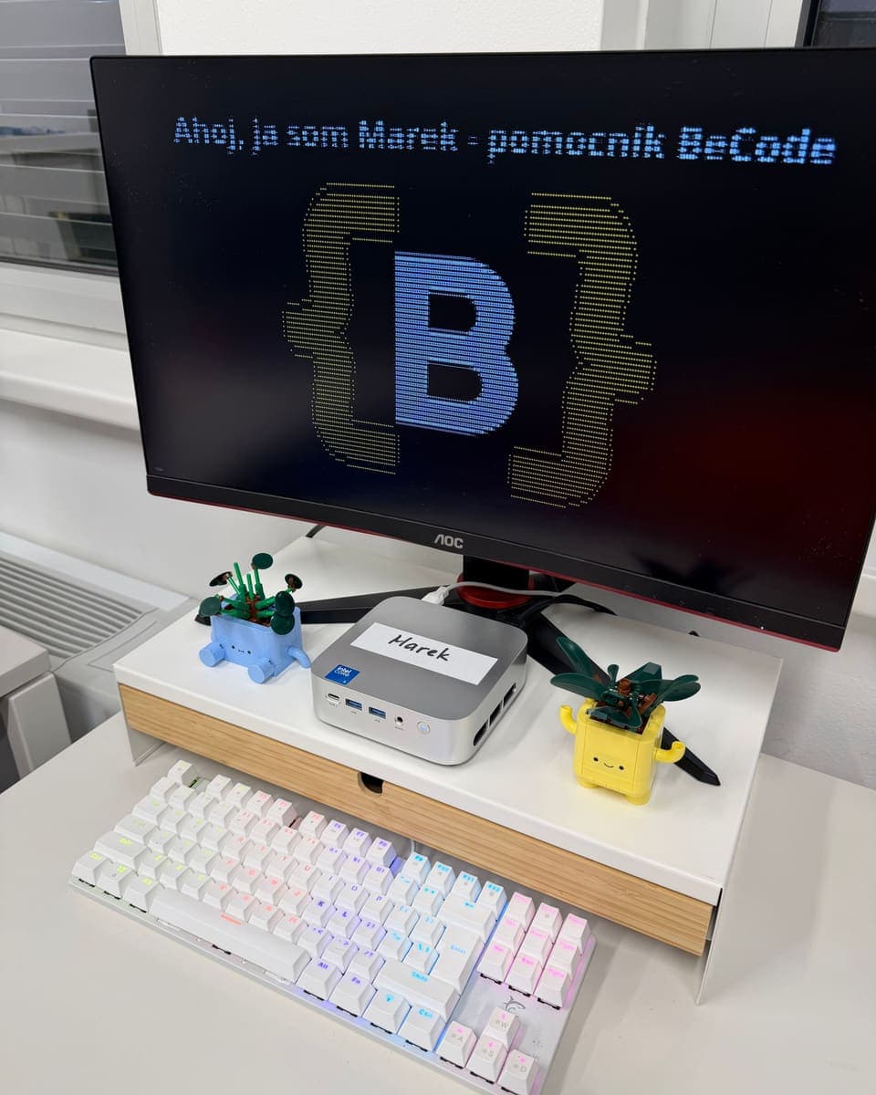 Marek v naší kanceláři: stříbrné mini PC se jmenovkou „Marek“ pod monitorem, na kterém svítí pozdrav „Ahoj, ja som Marek – pomocník BeCode“
